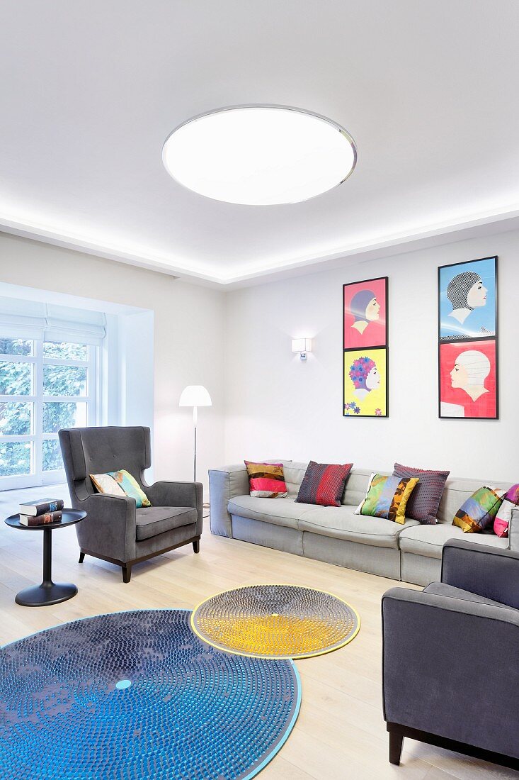 Bunte Kissen auf Sofa und Sessel in Grautönen, darüber Bilder im Pop-Art Stil und runde, farbige Teppiche auf dem Boden