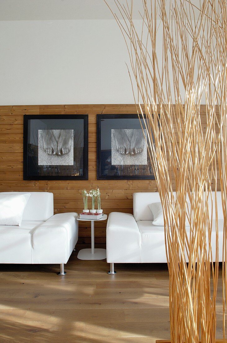 weiße Sofas vor schwarzweissen Kunstfotos an halbhoher Wandvertäfelung, davor ein Raumteiler aus Reisigbündeln