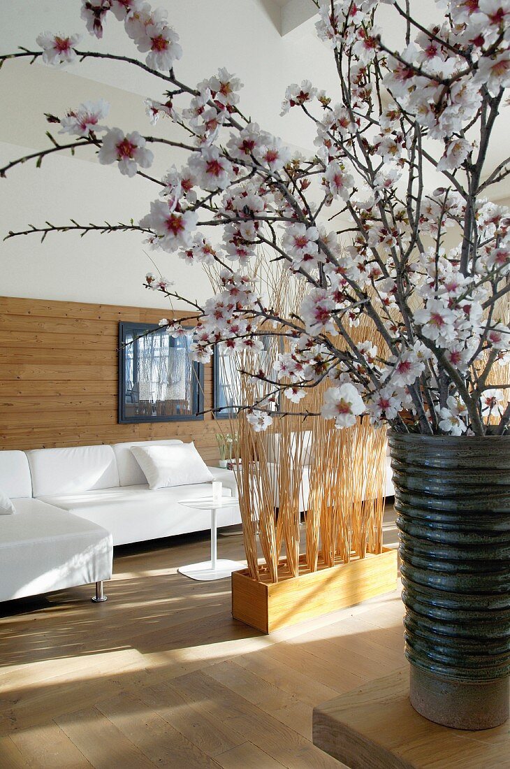 Halbhohe Wandvertäfelung hinter weissen Sofas und Raumteiler aus Reisigbündeln; im Vordergrund eine Vase mit Kirschblütenzweigen