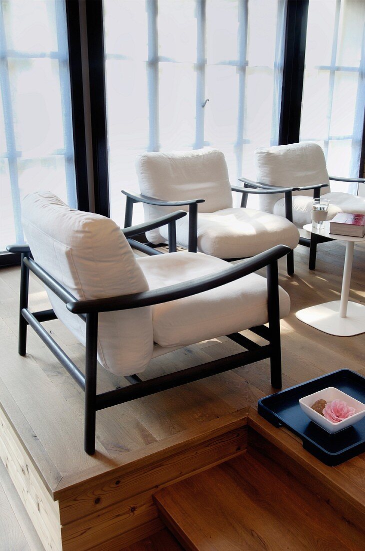 Schwarze, elegante Sesselgestelle mit weissen Polstern und Tablett mit Blütenschale auf einem Holzpodest vor Fensterfront