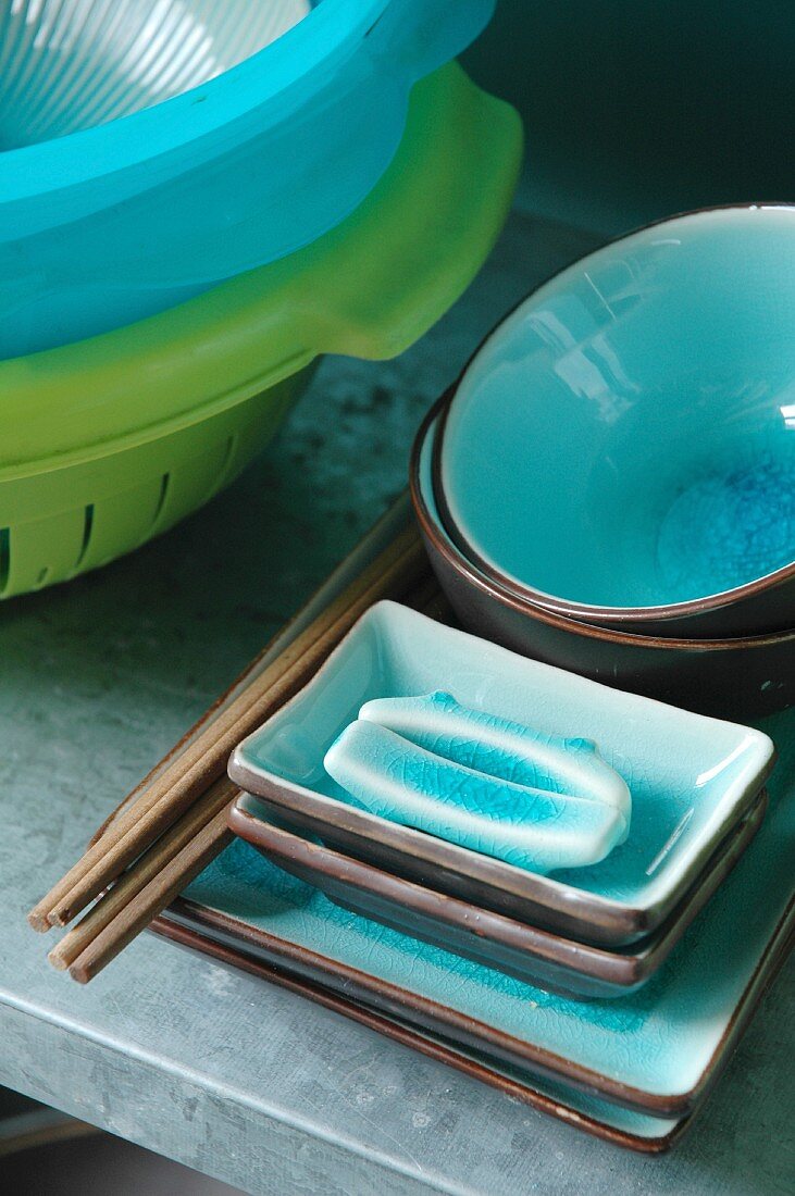 Chopsticks on stacked ceramic bowls with turquoise-glazed insides and brightly coloured plastic colanders on steel shelf in kitchen