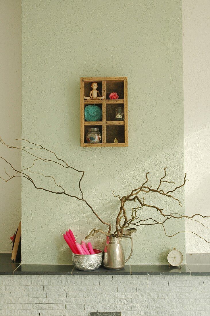 Silver jug of branches and dish of candles on mantelpiece below small, old display case on wall