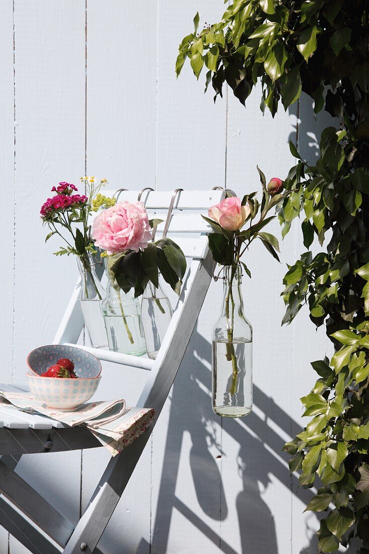 Bottles of flowers hanging on garden chair against white wooden wall