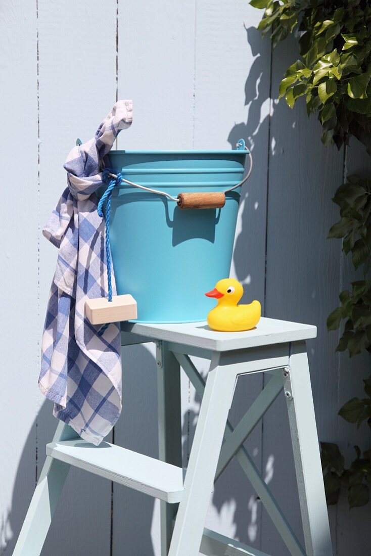Blue metal bucket, bar of soap, cloth and yellow rubber duck on white wooden step ladder