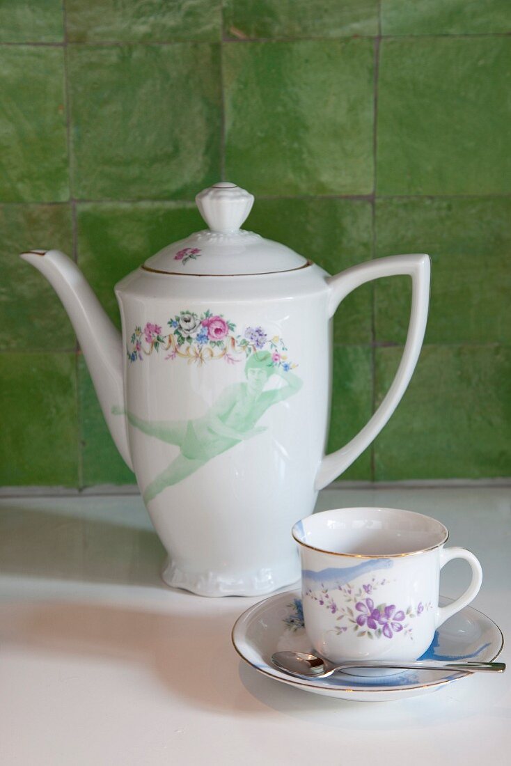 Coffee pot, cup and saucer with floral pattern in front of green wall tiles