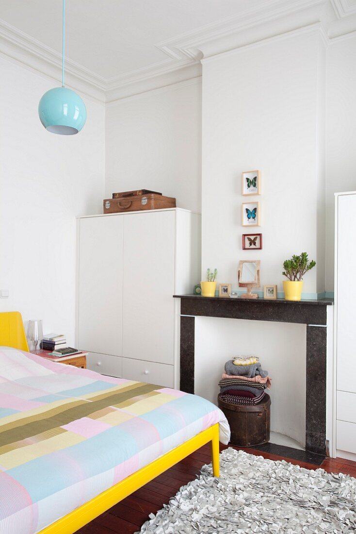 Bed with yellow frame and patchwork bed linen next to disused fireplace between white cupboards