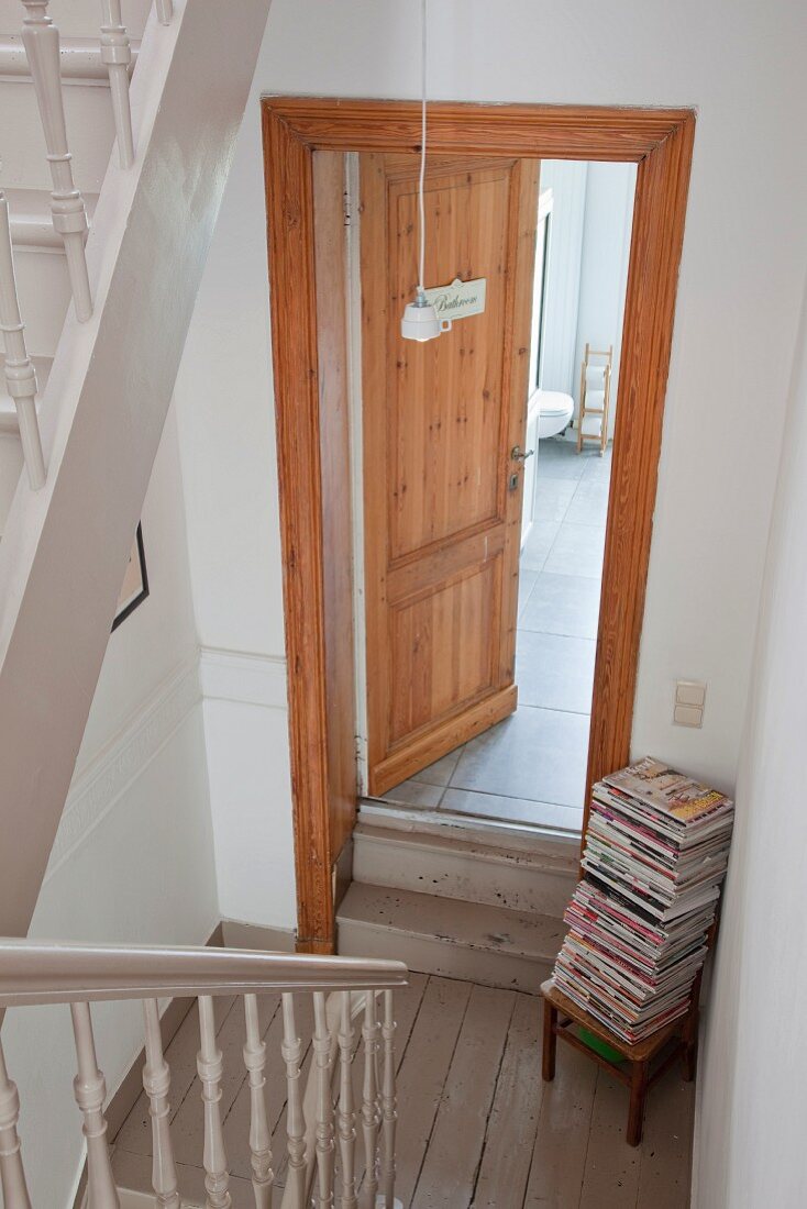 View down stairwell to open bathroom door and stack of magazines on chair in corner of landing