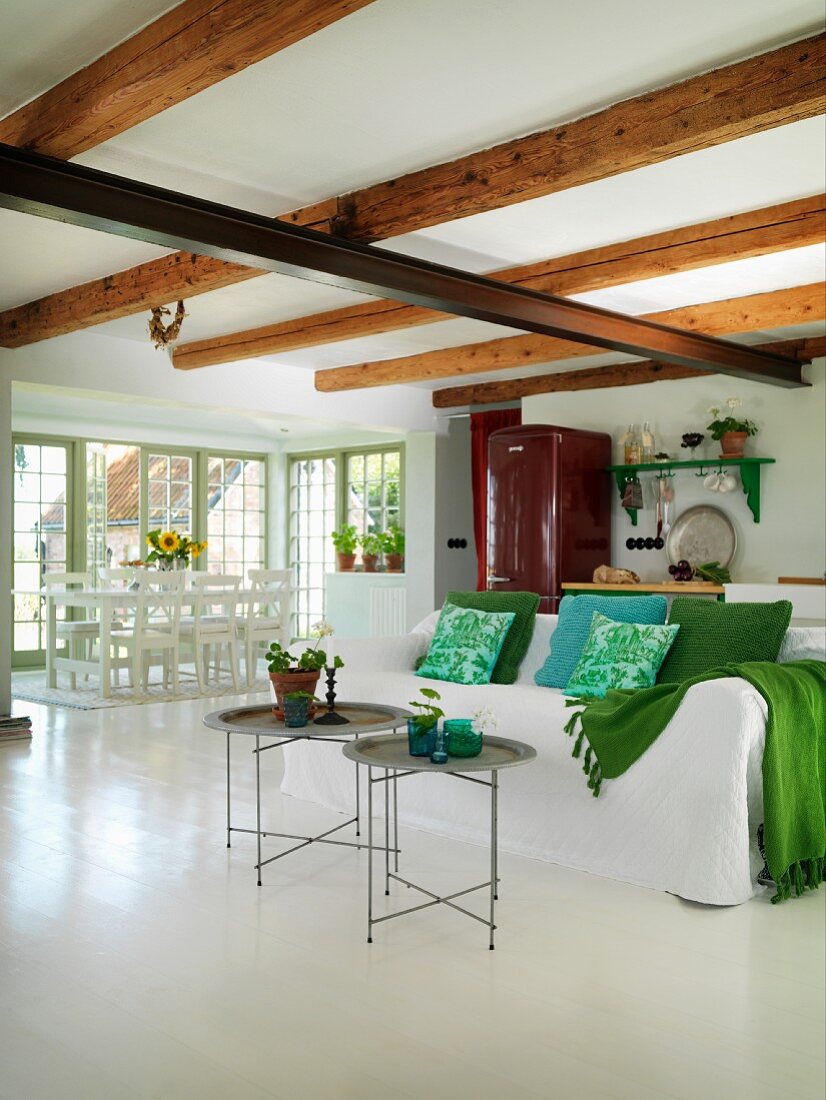 Delicate tray tables in front of sofa with white loose cover and scatter cushions in various shades of green in open-plan interior with dining area in background