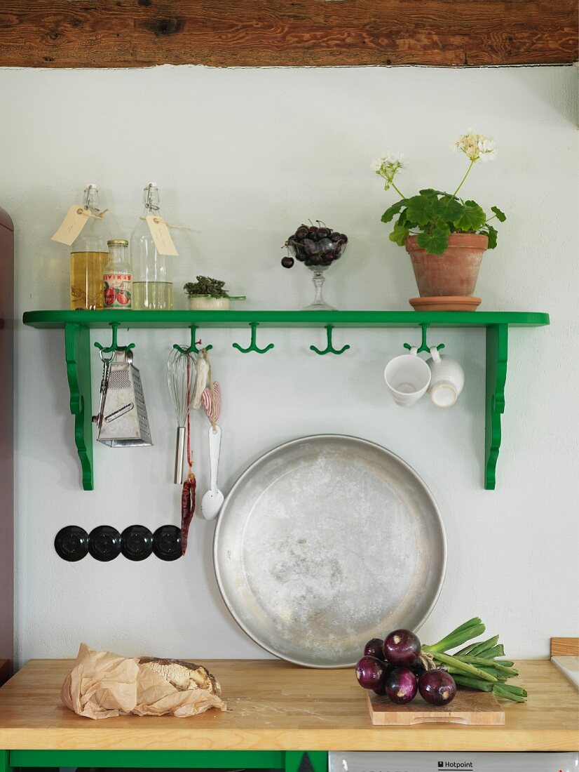 Green-painted wall-mounted shelf with hooks above metal tray leaning against wall on wooden kitchen worksurface