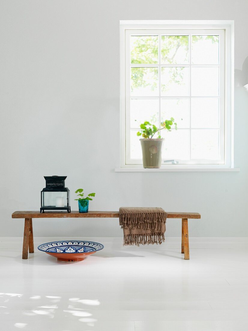 Rustikale Holzbank mit Laterne und Keramikschale auf weißem Holzdielenboden vor Sprossenfenster