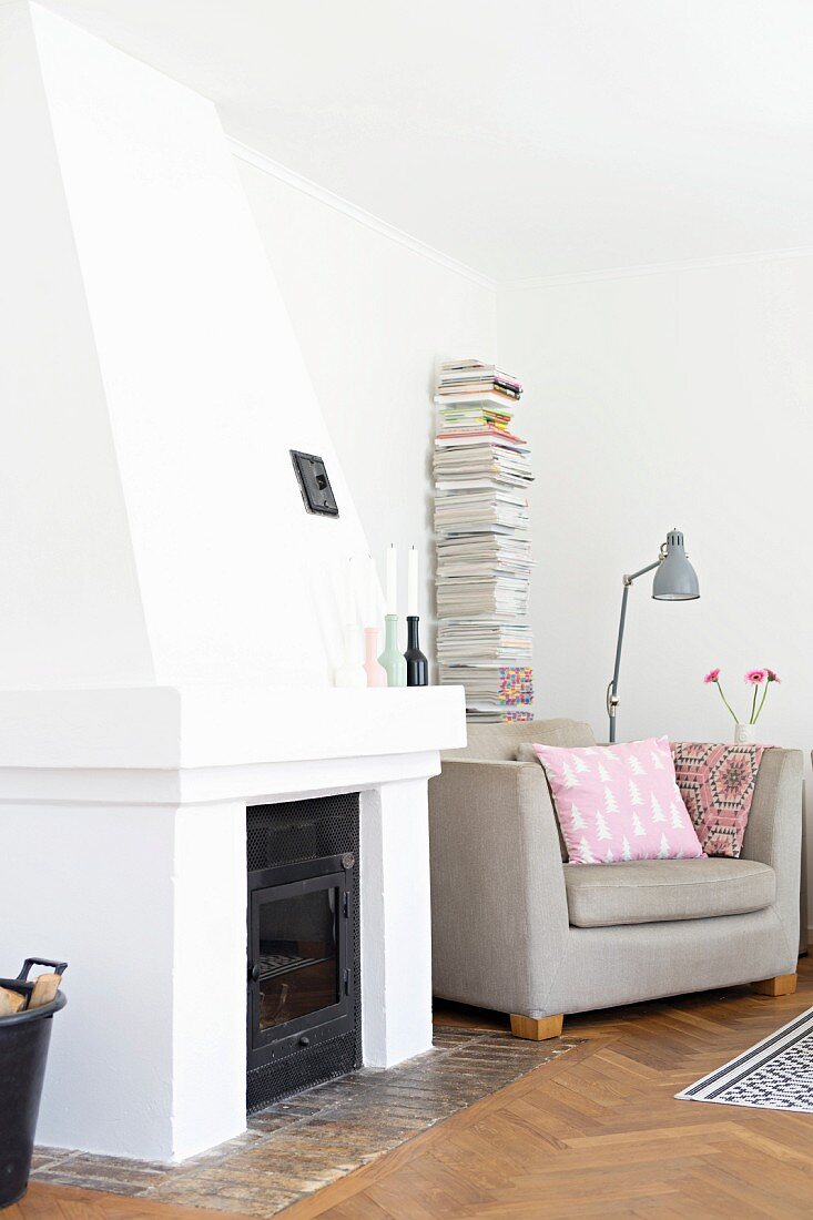 Wohnzimmerecke mit offenem Kamin, seitlich naturfarbener Polstersessel, dahinter Zeitungsstapel in minimalistischem Regal vor Wand