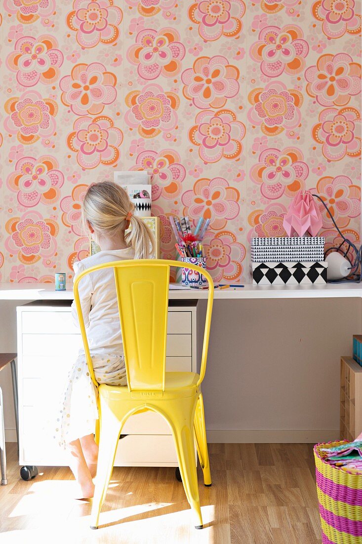 Mädchen auf gelb lackiertem Metallstuhl vor Schreibtisch, an Wand Retro Blumentapete