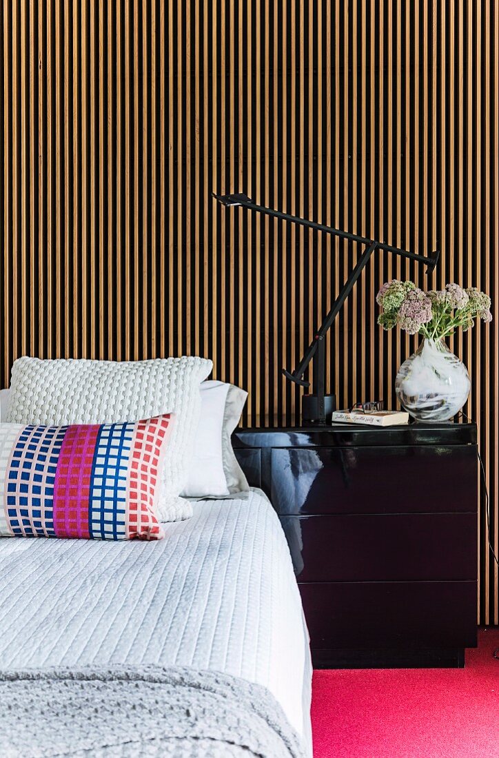 Kissen auf Bett mit Tagesdecke, seitlich schwarzer Nachttisch vor Wand mit Holzlamellenverkleidung