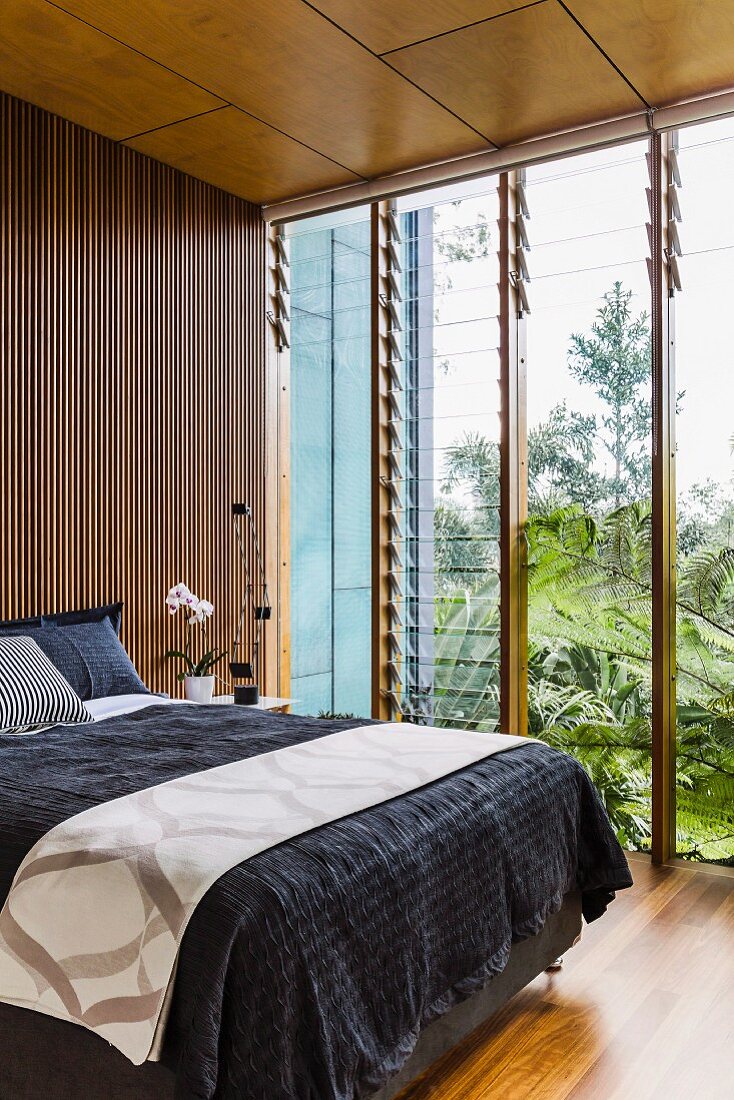 Doppelbett mit schwarzem Plaid vor Wand mit Holzlamellenverkleidung, seitlich Glasfront mit verstellbaren Lamellen