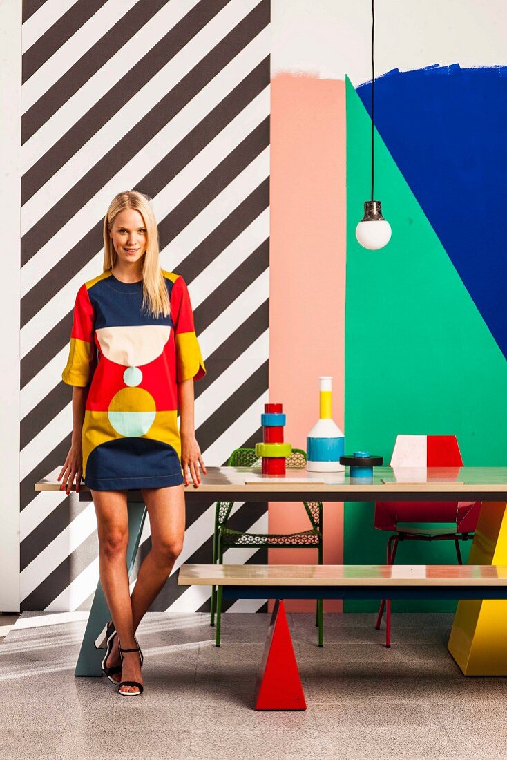 Young woman wearing retro mini-dress in front of colourful graphic pattern on wall and still-life arrangement of vases on table