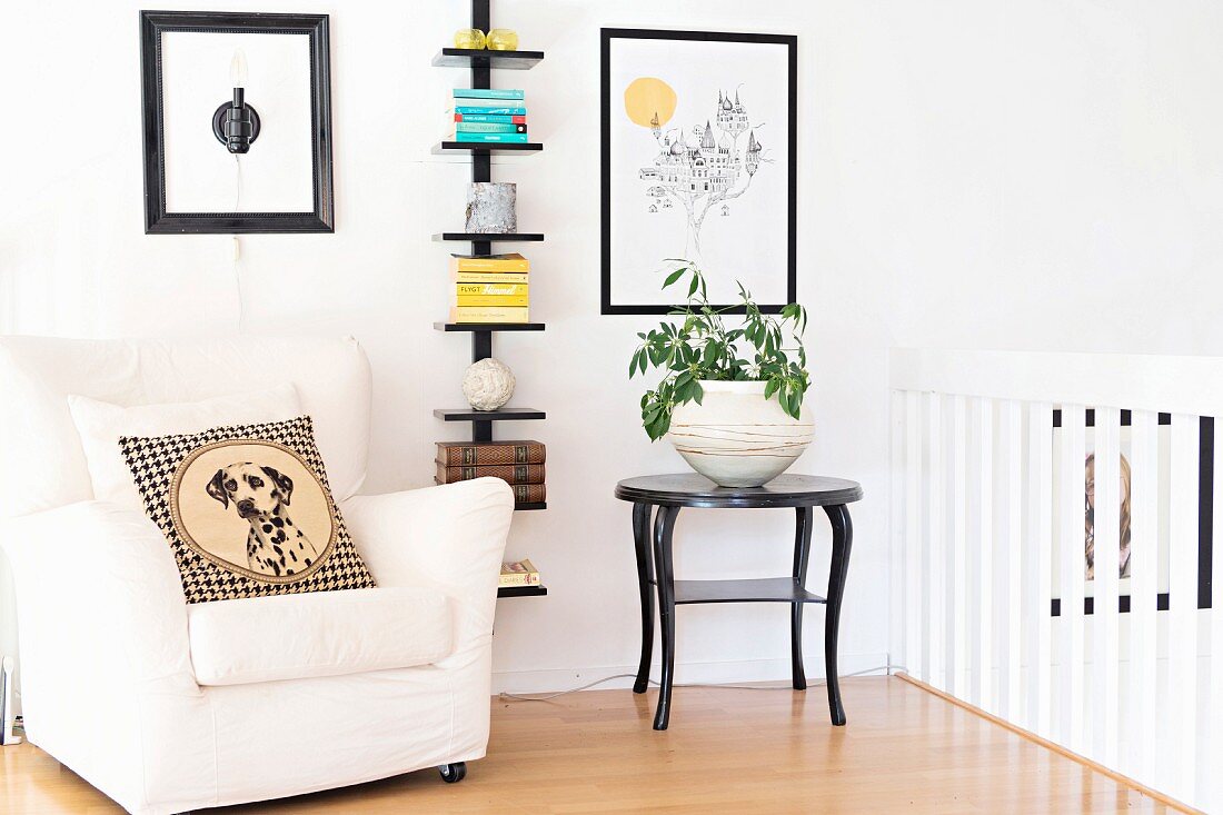Cushion with dog motif on white armchair, potted plant on black side table and stacked books on wall-mounted shelves
