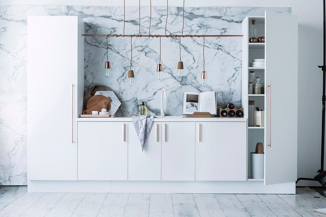 weiße Miniküche mit Hochschränken und integrierter Küchenzeile, oberhalb um Stange gewickelte Pendelleuchten in Reihe, Wandgestaltung mit Marmor Effekt