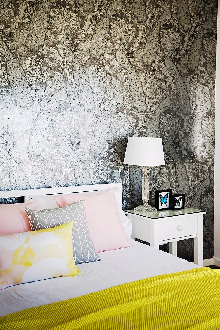Weisses Bett mit gelbem Überwurf und Pastell Kissen vor gemusterter Tapetenwand mit Pfauenmotiv in Grautönen