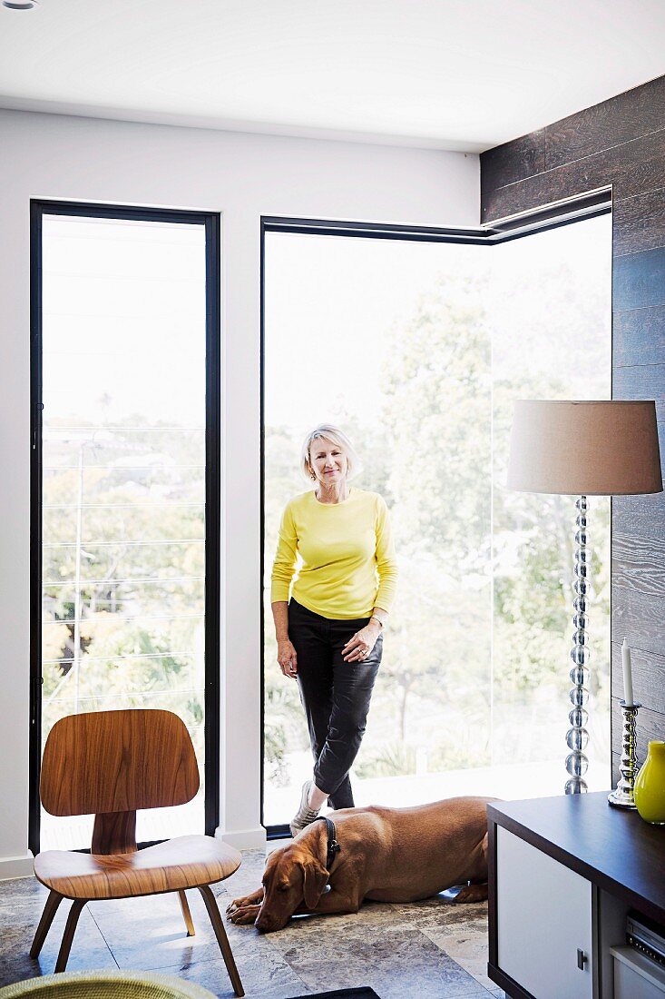 Woman wearing yellow top and dog in front of frameless corner window, replica of classic chairs and postmodern standard lamp with plexiglas base