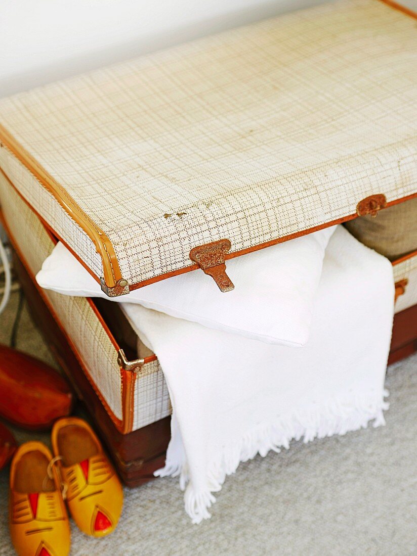 Pale vintage suitcase, white blanket and wooden clogs arranged on carpeted floor