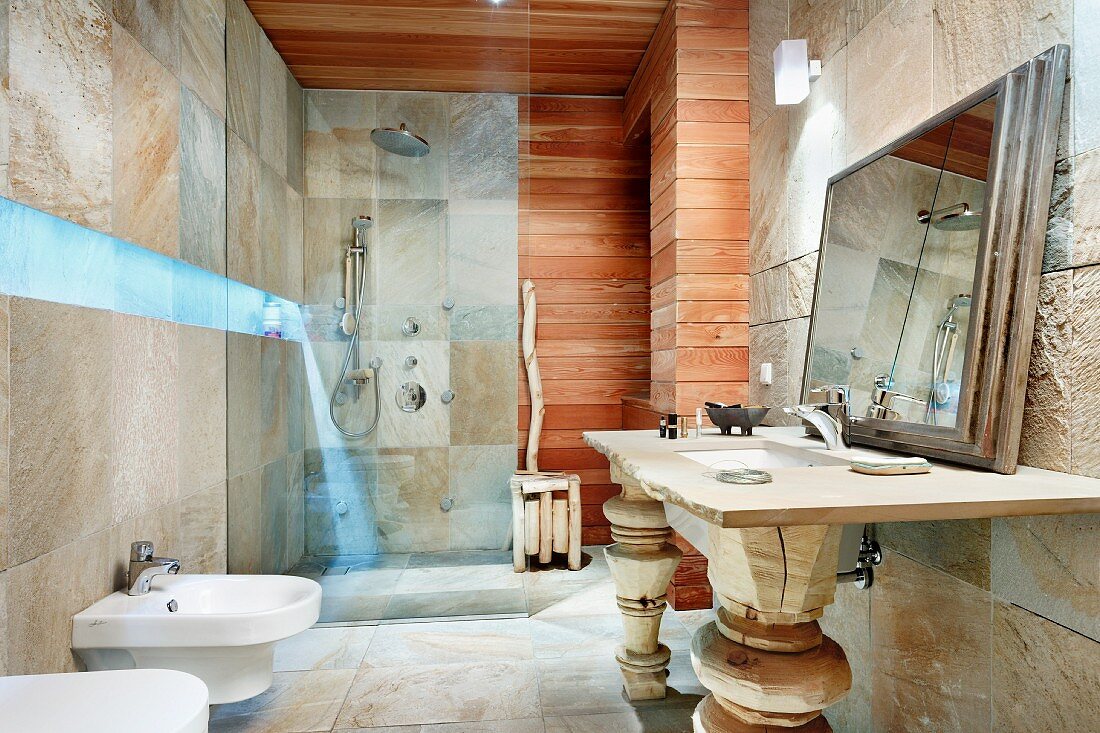 Bad mit bläulichem Leuchtband zwischen Natursteinfliesen und horizontaler Holzverkleidung, Waschtisch mit geschnitzten Holzstützen und gerahmtem Spiegel