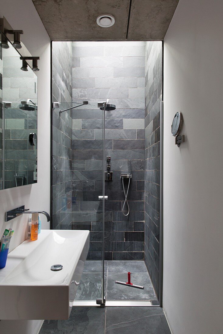 Schmales, modernes Bad, seitlich Waschbecken mit Wandarmatur, im Hintergrund bodenebener Duschbereich mit gefliesten Wänden aus Schieferplatten
