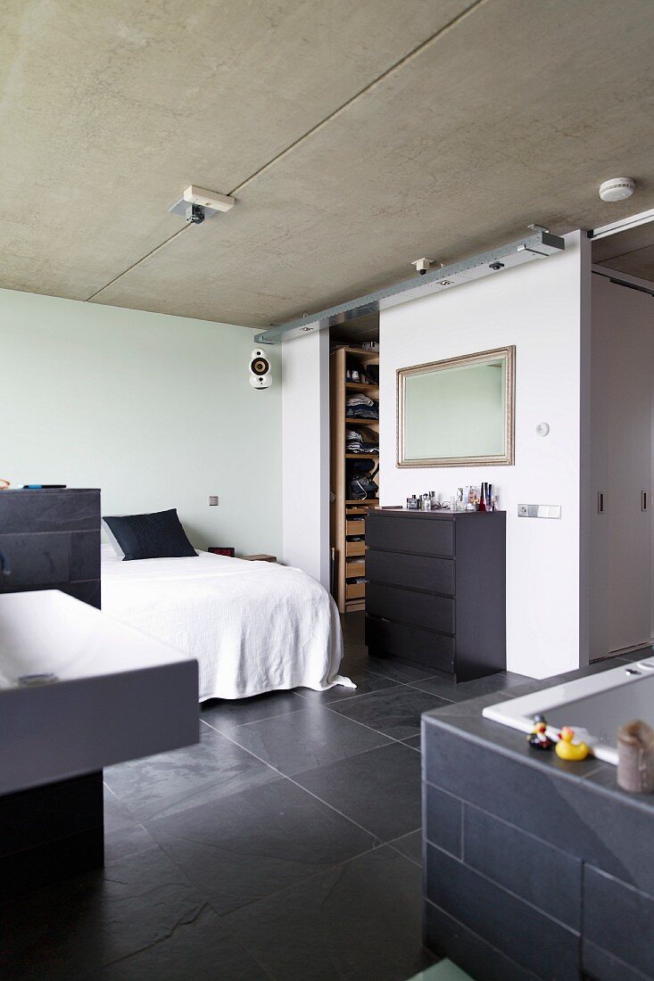 Schlafzimmer mit Sichtbetondecke und Schieferboden, im Vordergrund teilweise sichtbarer, offener Badbereich