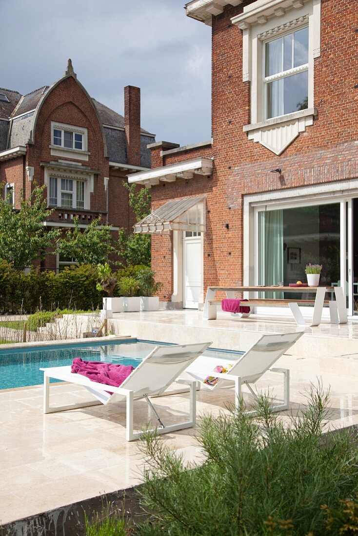 Sun loungers on side of modern pool outside traditional Belgian brick house with white decorative elements around windows