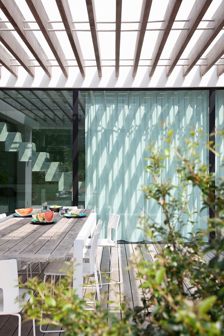 Wooden terrace below slatted pergola adjoining modern house with pattern of light and shadow on glass façade