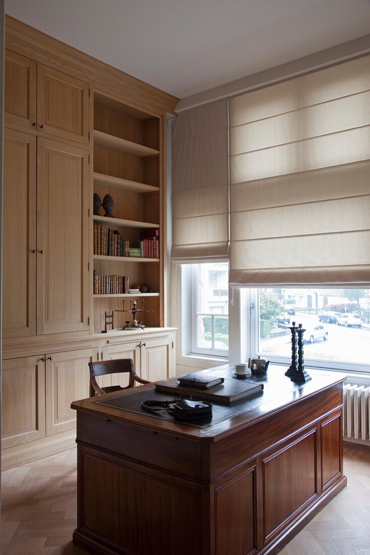 Solid wood desk in front of wooden fitted cupboards next to windows with half-closed roller binds