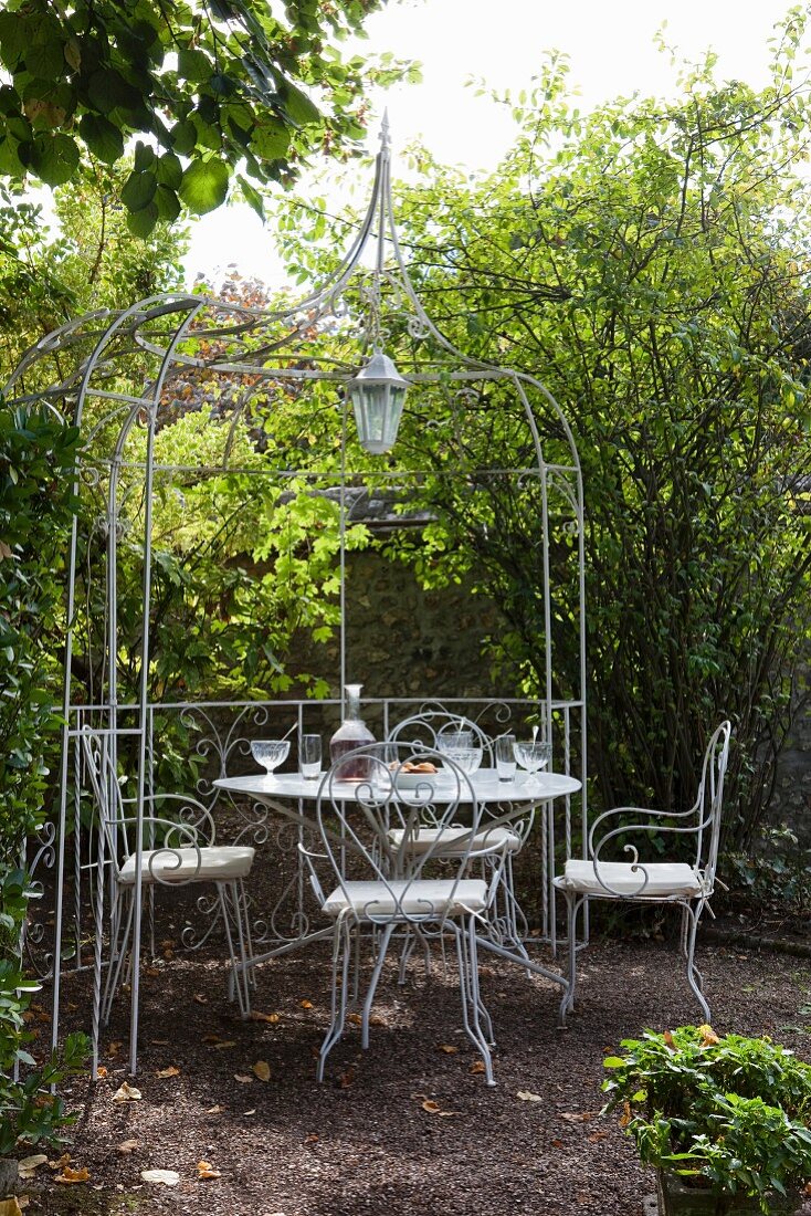 Filigrane Metallstühle und Tisch unter pavillonartiger Pergola aus Metall in idyllischem Garten