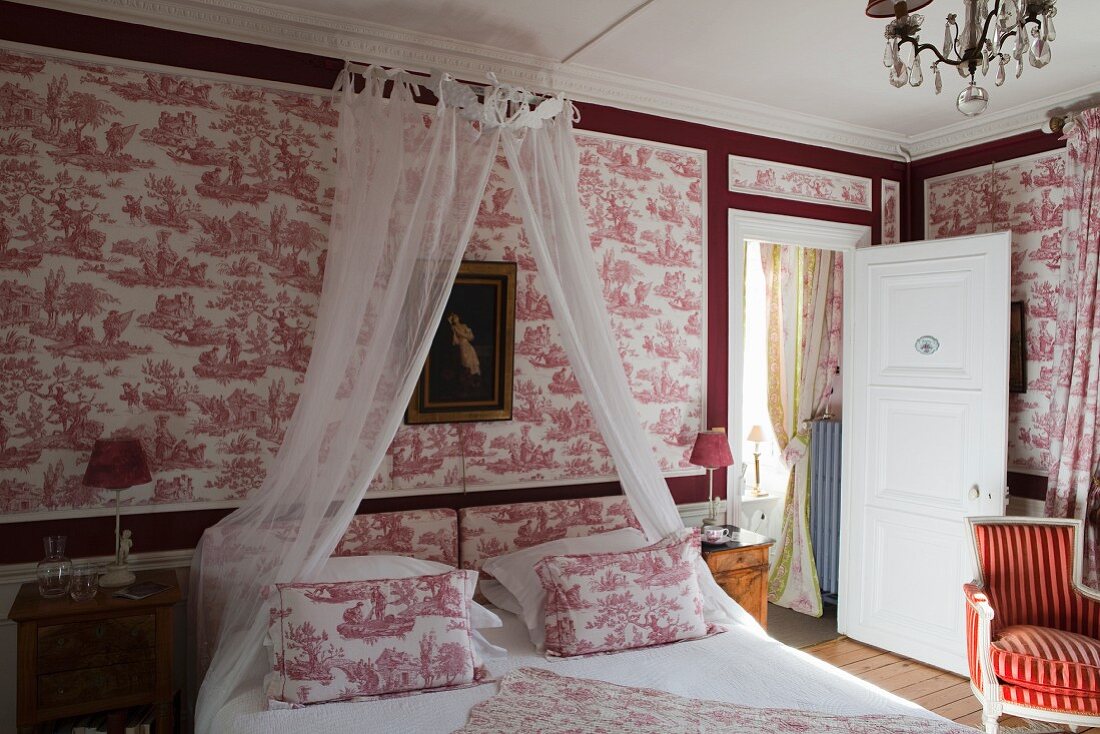 Double bed with canopy against red and white toile de jouy wallpaper