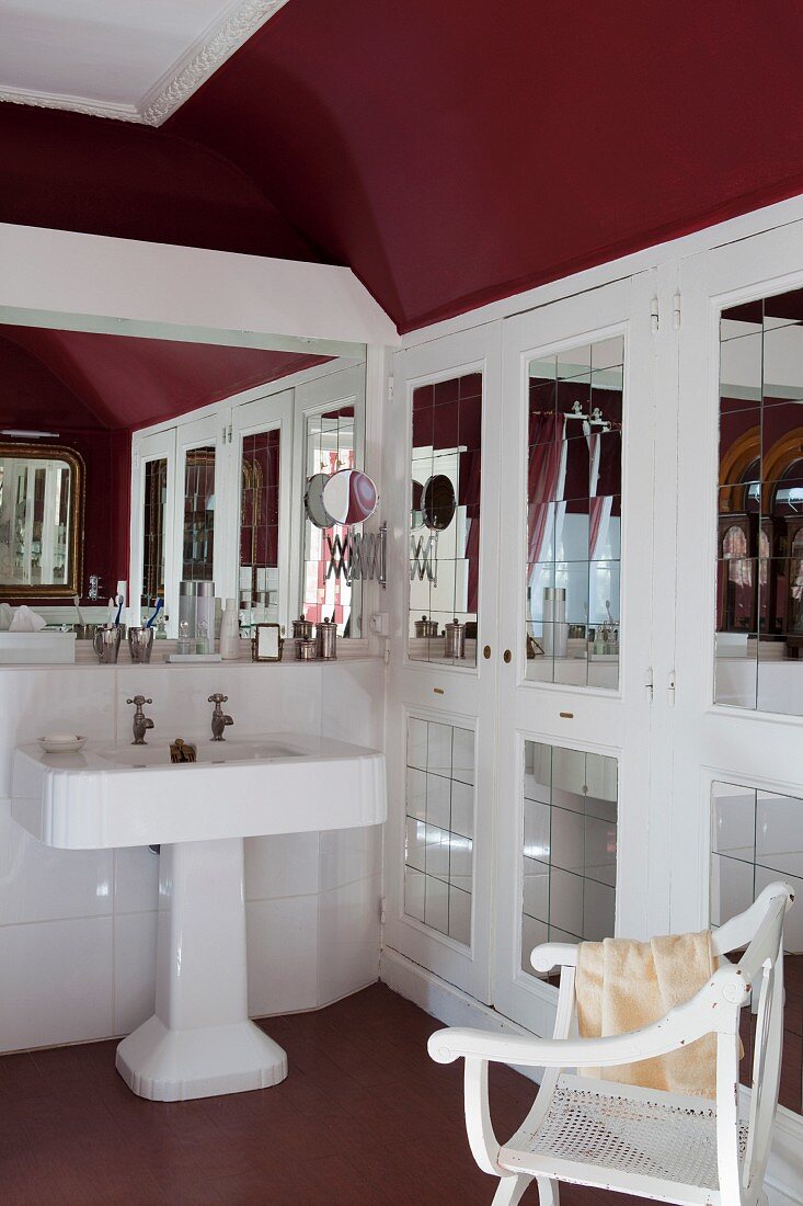 Vintage pedestal sink next to fitted cupboards with mirrored door panels and armchair in bathroom with dark red walls