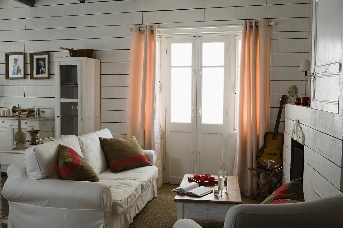 Sofa with scatter cushions next to French windows with curtains in corner of living room with white wood-clad walls