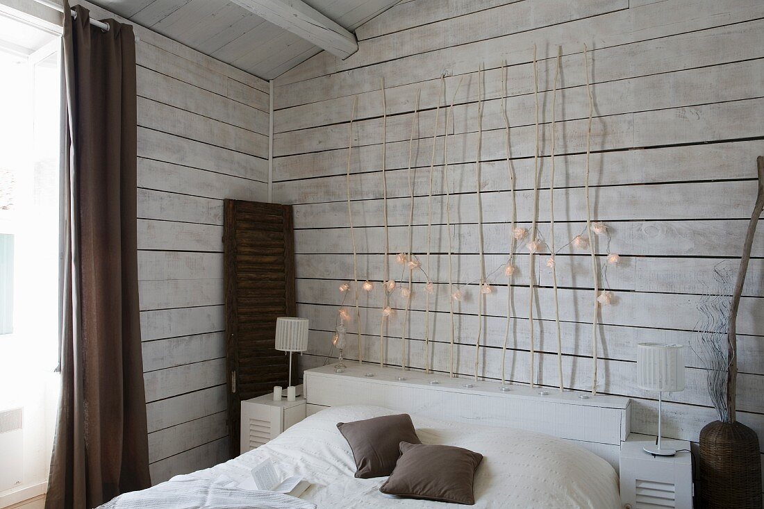 Simple bed with wooden headboard made of branches decorated with fairy lights against white, wood-clad wall
