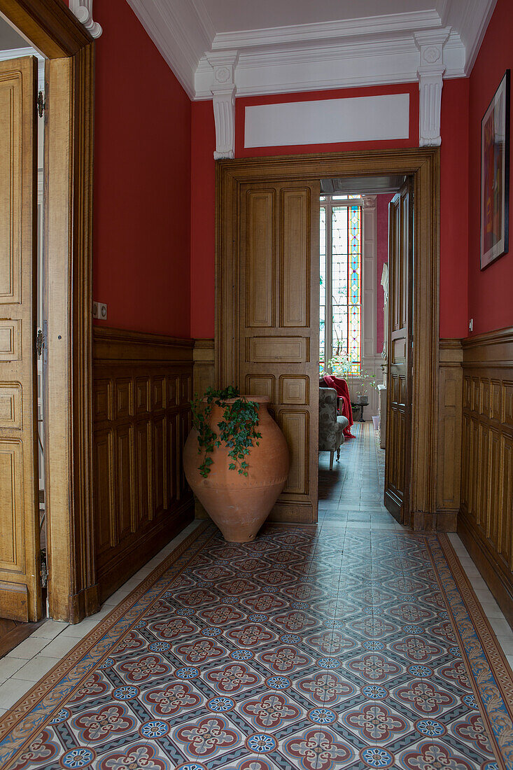 Ornamental tiled floor, plain wooden wainscoting, red-painted walls and amphora next to open double doors in elegant hallway of grand villa