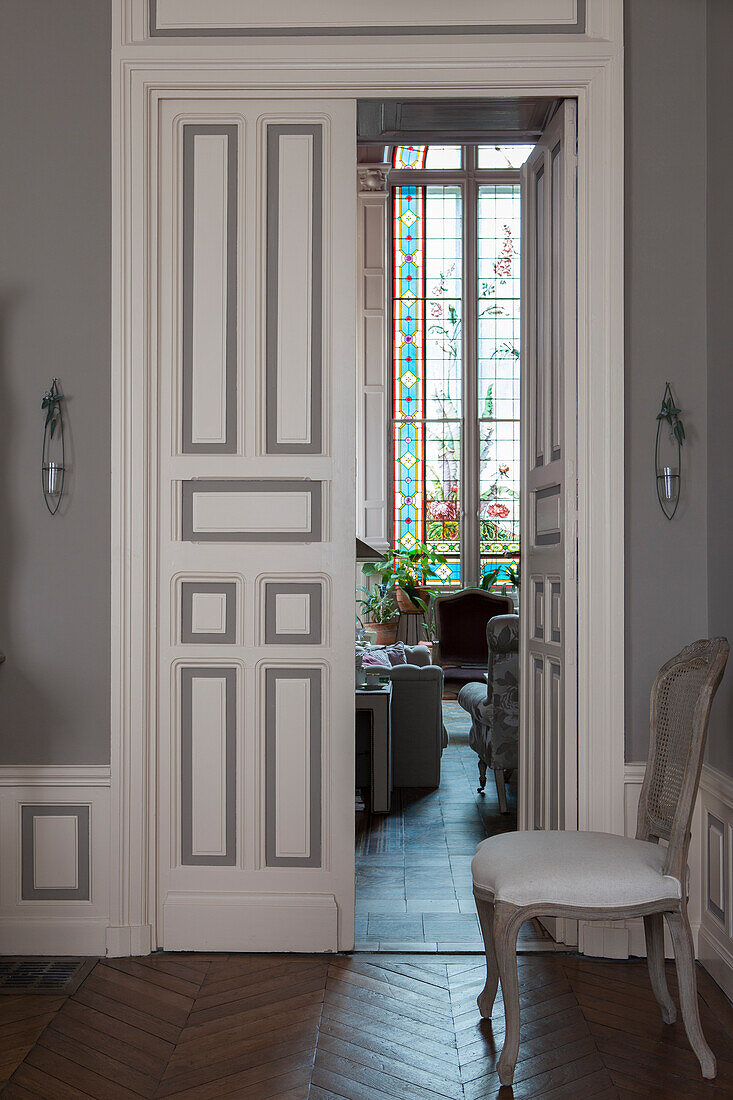 Cane-backed chair with upholstered seat next to open door and view of floor-to-ceiling window with artistic stained-glass elements