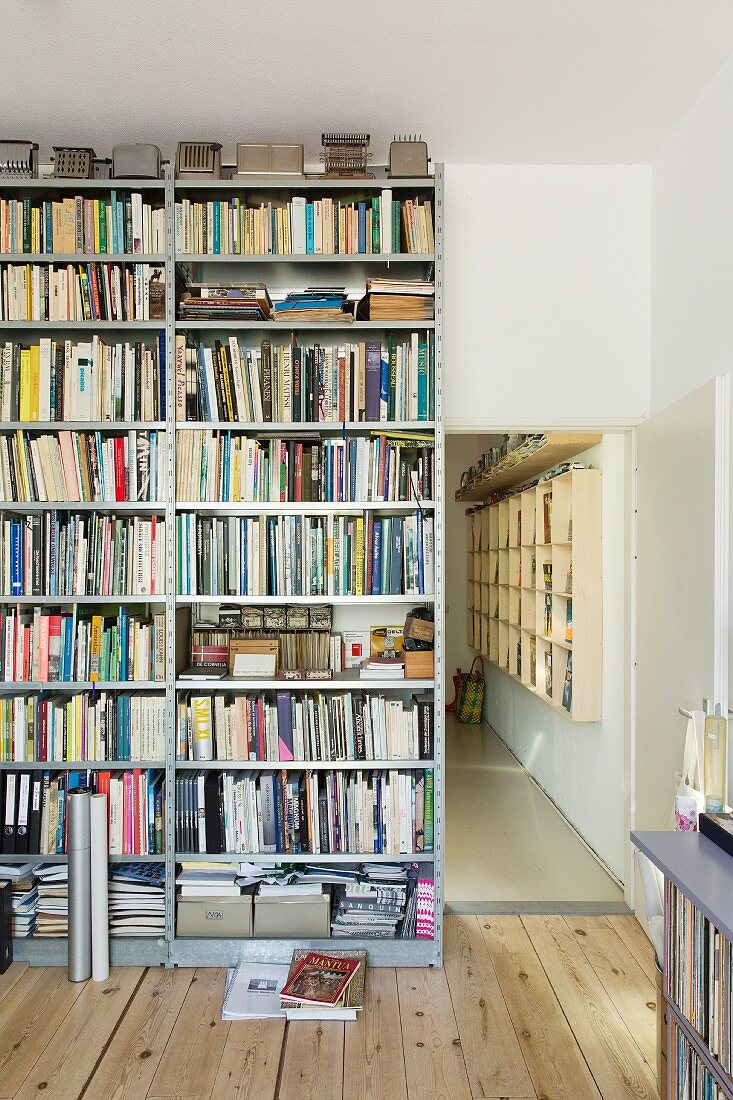 Floor-to-ceiling metal bookcase next to open doorway with view of wooden wall-mounted shelves in hallway