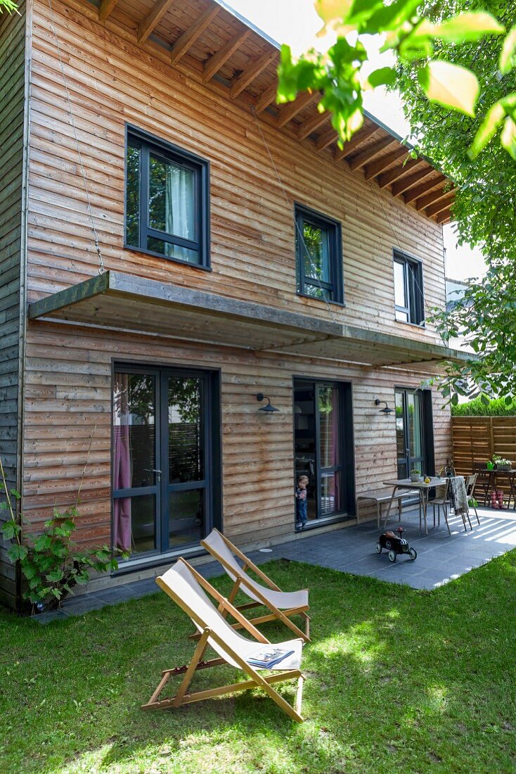Liegestühle im Garten vor zeitgenössischem Holzhaus, kleine Terrasse mit Sonnenschutz