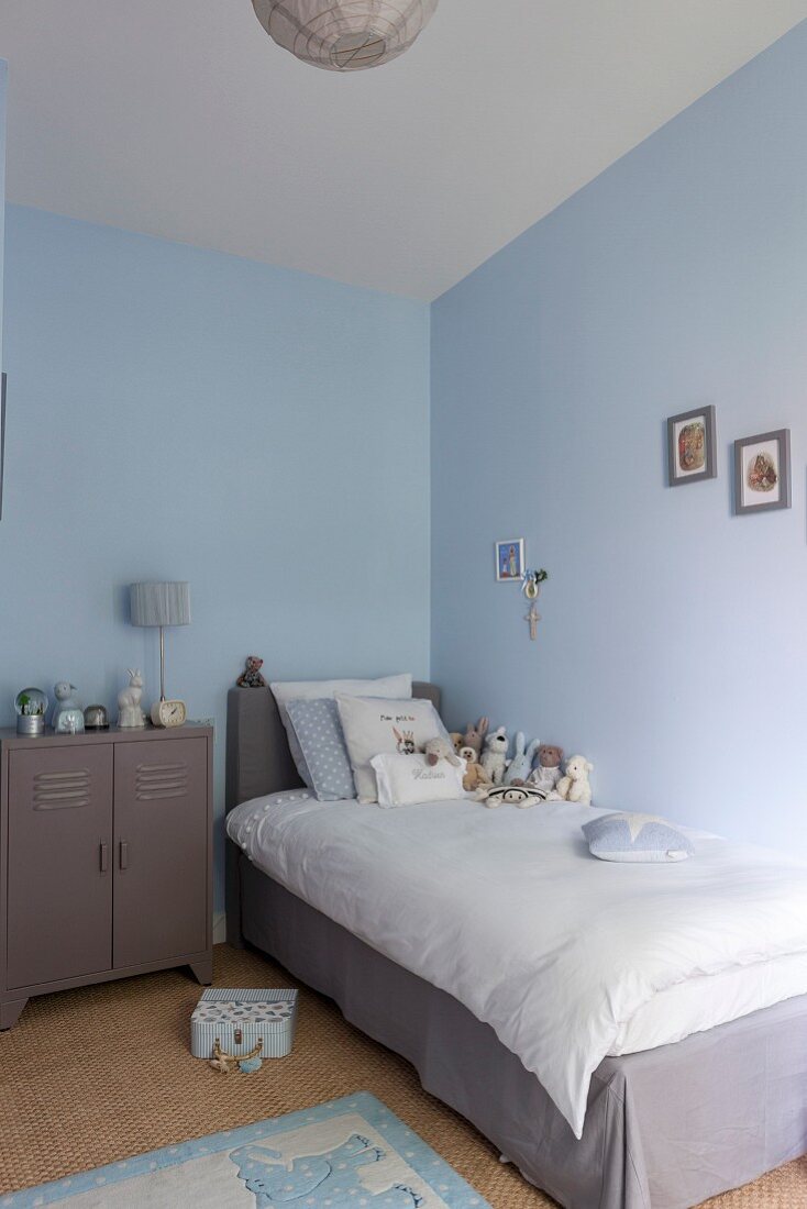 Einzelbett mit grauer Husse und weisser Bettwäsche, neben Retro Metall Schränkchen in Kinderzimmerecke mit hellblauen Wänden