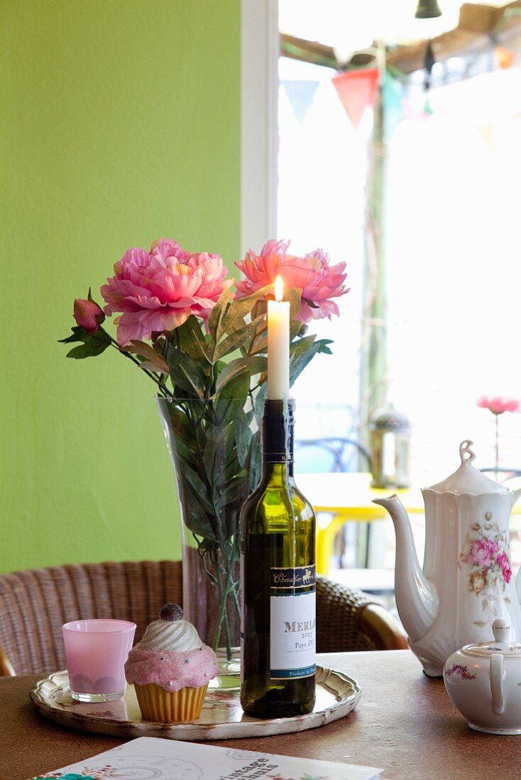 Lit candle in bottle in front of vase of peonies on tray next to floral coffee pot