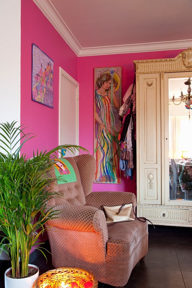 Polstersessel und Zimmerpalme vor pinkfarbener Wand, im Hintergrund antiker Schrank mit Spiegel an Tür