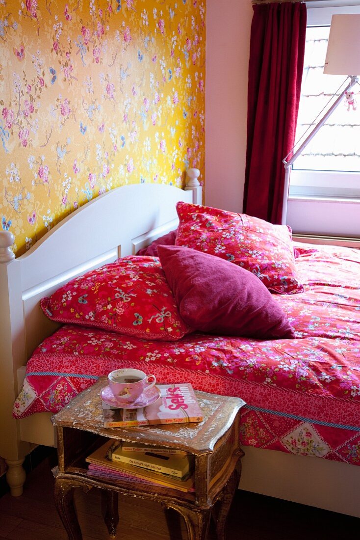 Red floral bed linen on double bed with white headboard against floral wallpaper