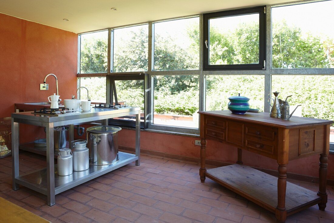 Kunsthandwerkliche Anrichte und Edelstahlspüle mit Milchkannen und Kochtopf in spärlich möbliertem Raum mit Fensterfront
