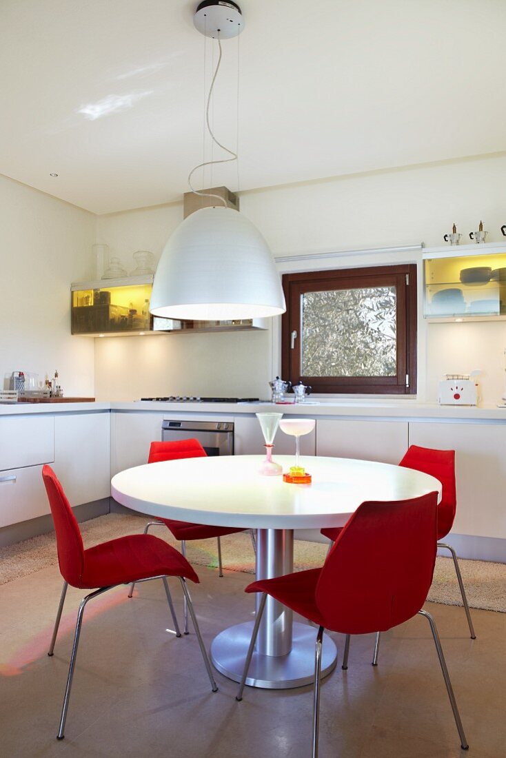 Weißer runder Tisch mit roten Designerstühlen in weißer Einbauküche mit gelb beleuchteten Hängeschränken