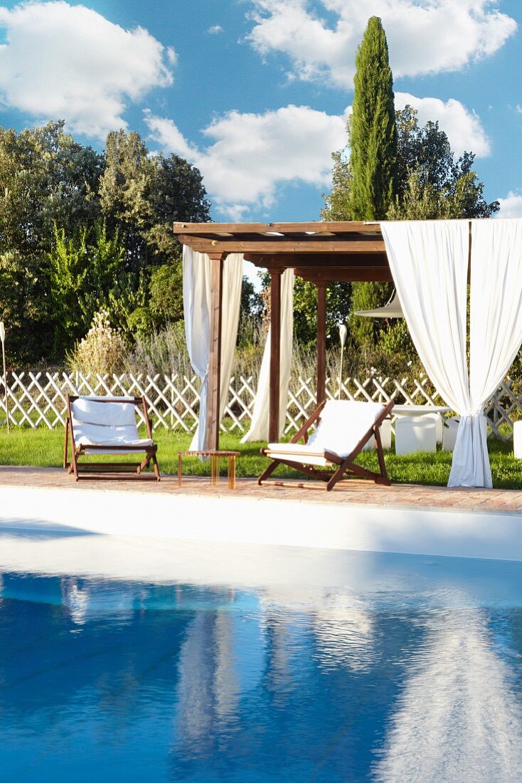 Holzpergola mit weißen Vorhängen und Holzliegestühle mit weißen Polstern im sommerlichen Garten am Pool