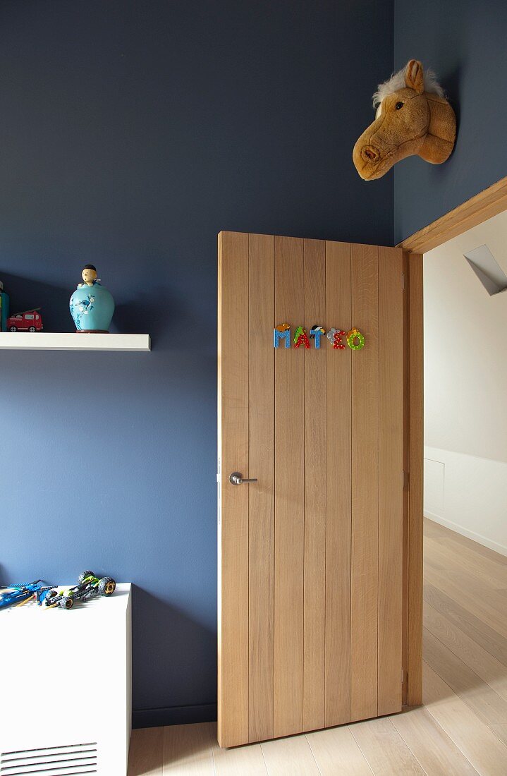 Jungenzimmer mit geöffneter Zimmertür aus hellem Holz und Stofftierkopf an blauer Wand