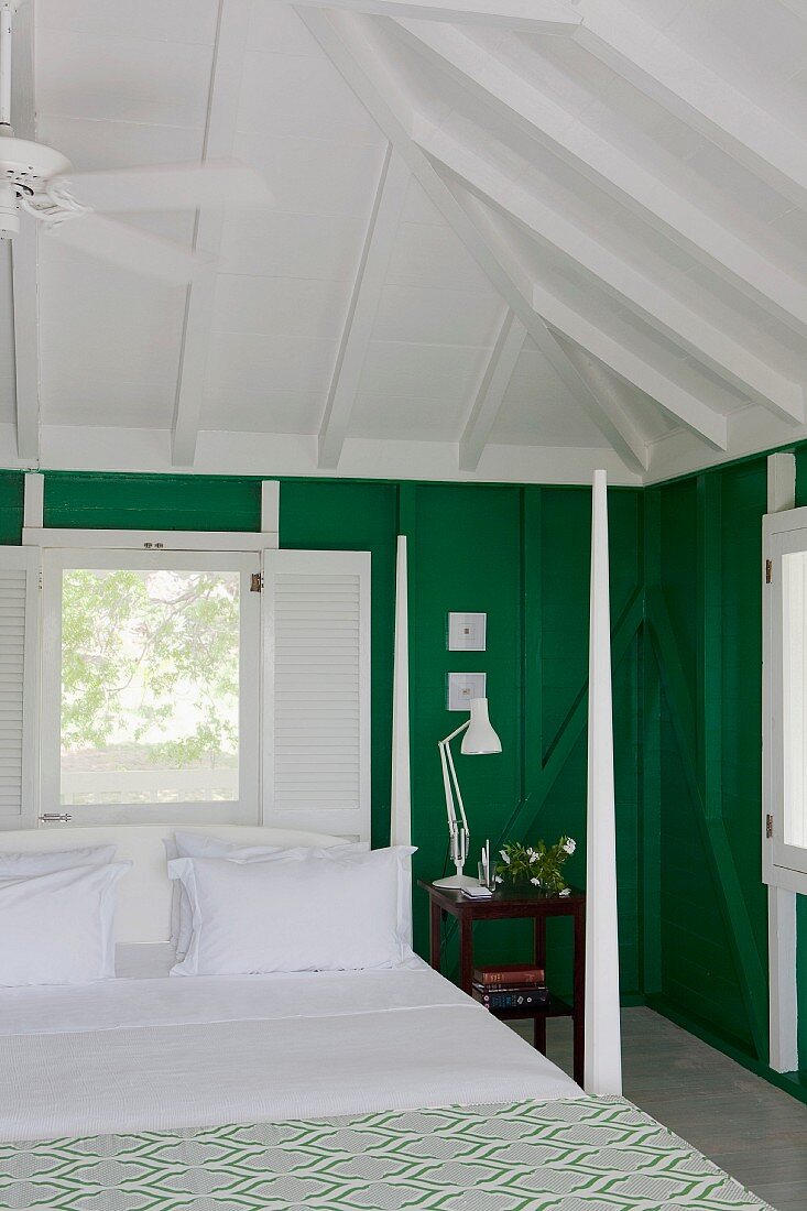 Bed with corner posts and snow-white bed linen below exposed pavilion roof structure, window with interior shutters and green-painted wooden walls