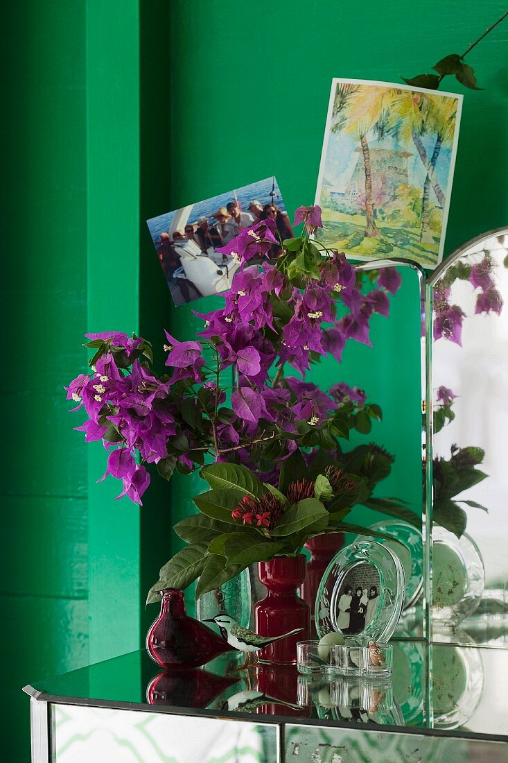 Erinnerungsstücke und Vase mit Bougainvillea auf einem antiken Spiegeltisch vor grüner Wand