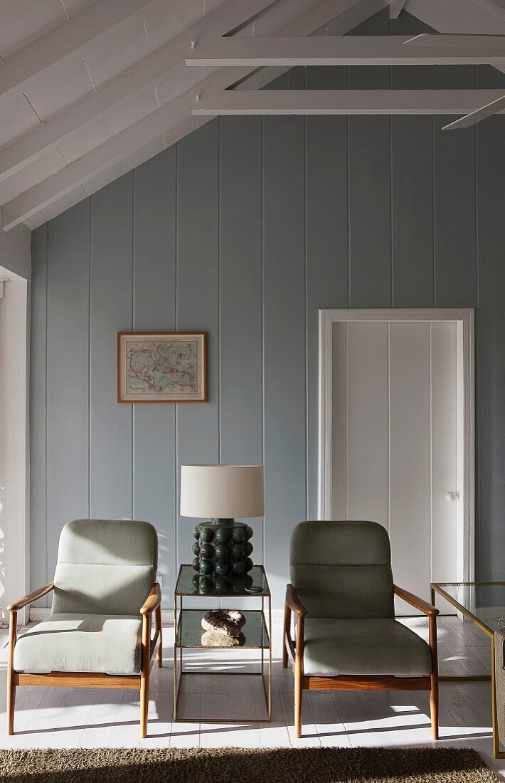 Lamp on mirrored table flanked by 50s retro armchairs in beach house painted grey and white with exposed roof structure